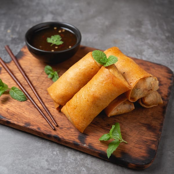 Golden-brown Thai spring rolls with mint garnish, dipping sauce, and chopsticks on a wooden board.