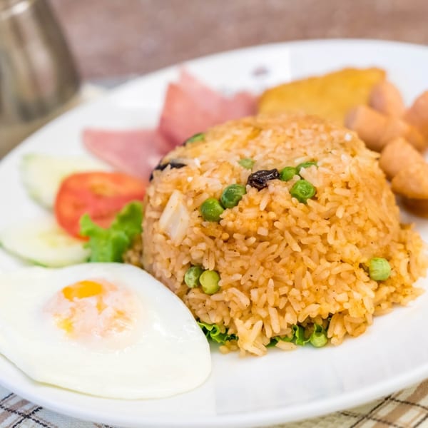 Thai fried rice with peas, ham, sausages, fried egg, and fresh vegetables on a white plate.