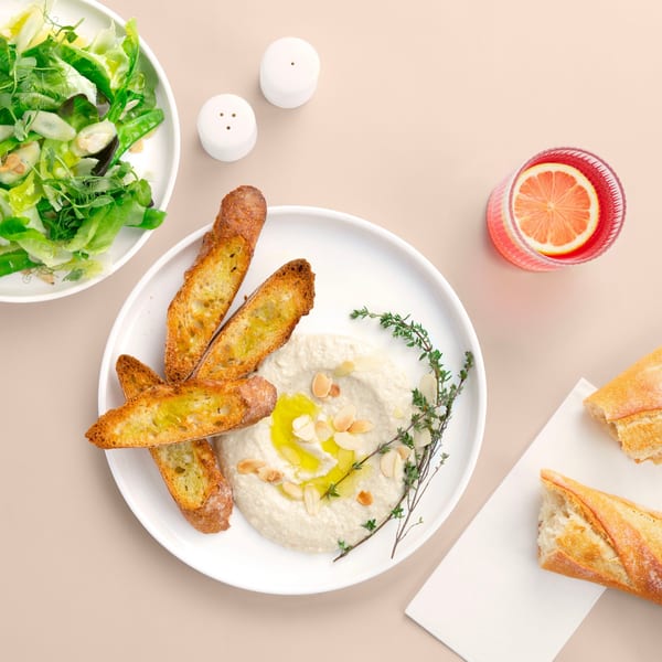 Overhead view of a Thai appetizer spread with hummus, toasted bread, green salad, and a pink drink.